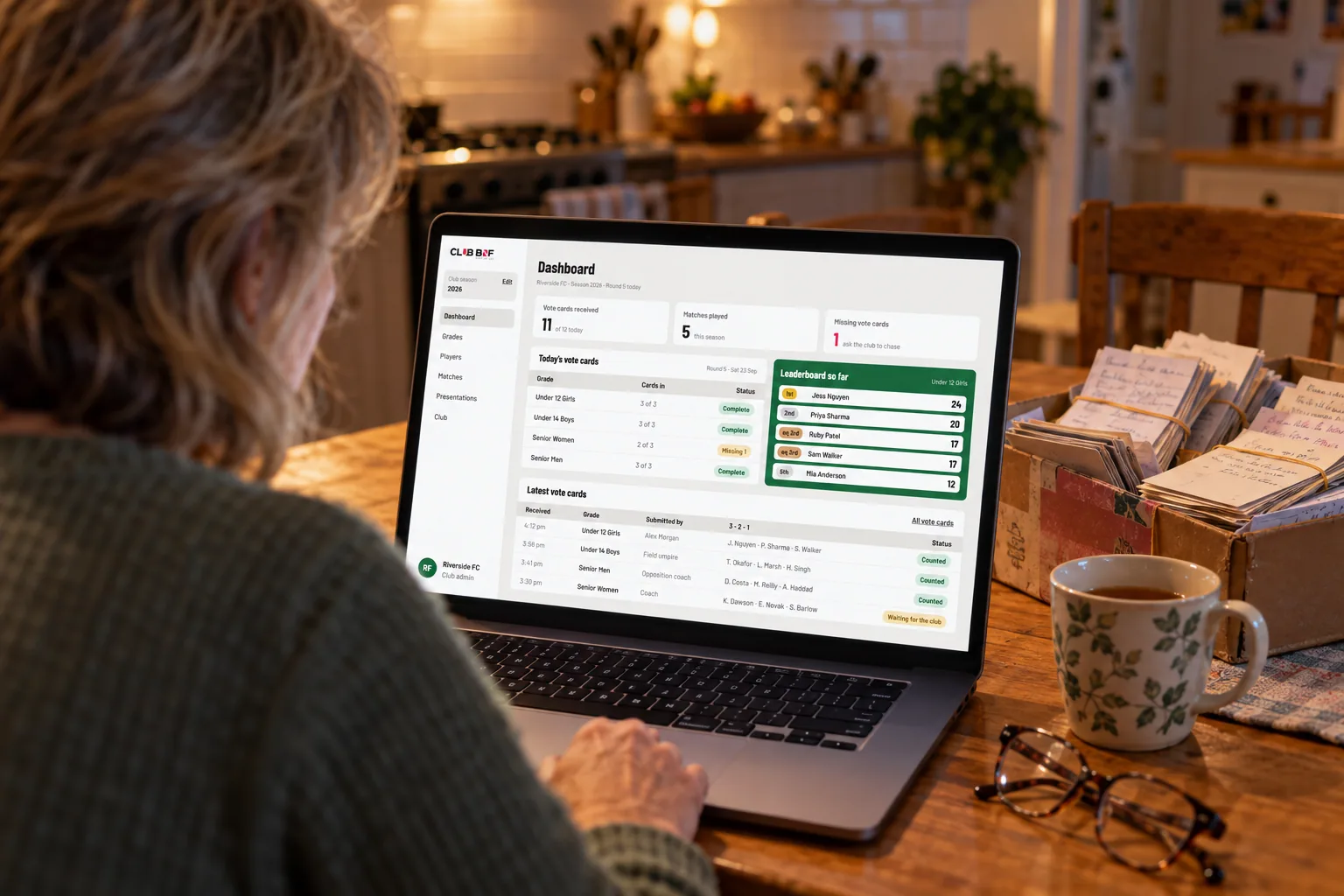 A club secretary at a kitchen table, a laptop open on the Club BNF dashboard beside a shoebox of old paper vote cards